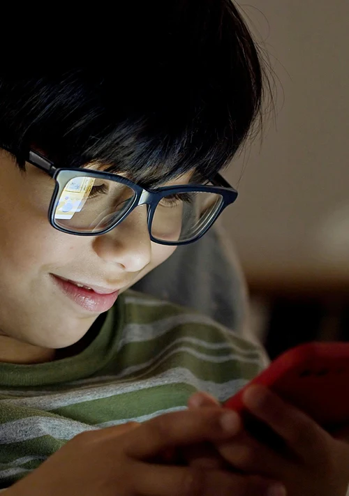 A smiling teen boy wearing glasses looks closely at his phone.