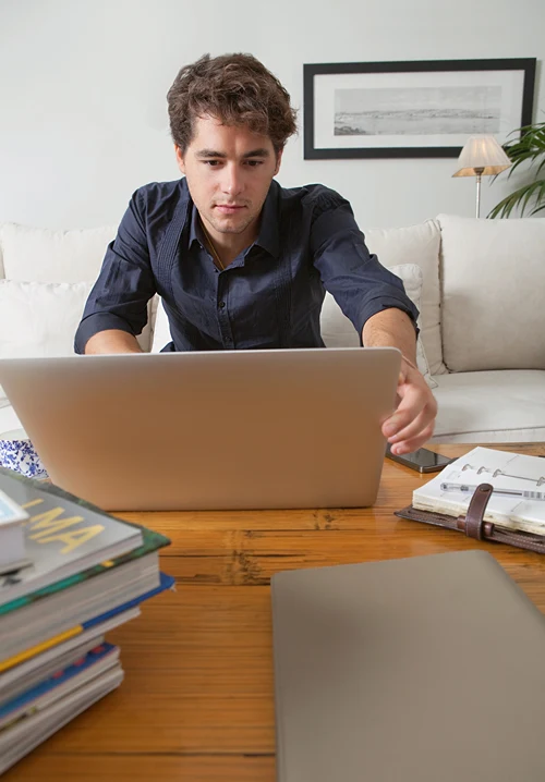 Male student surrounded by books while studying for an exam on his laptop.