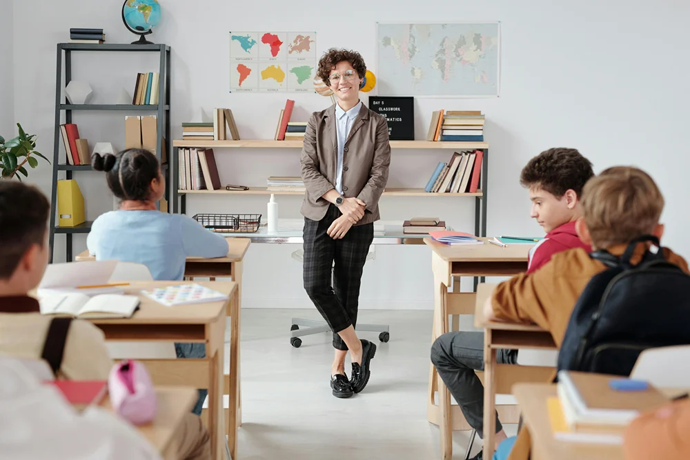 Smiling teacher leans against her desk in front of a classroom of students.