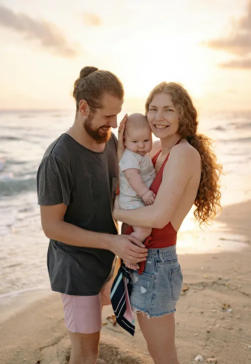 Woman holds her baby with husband standing on a sunny beach.