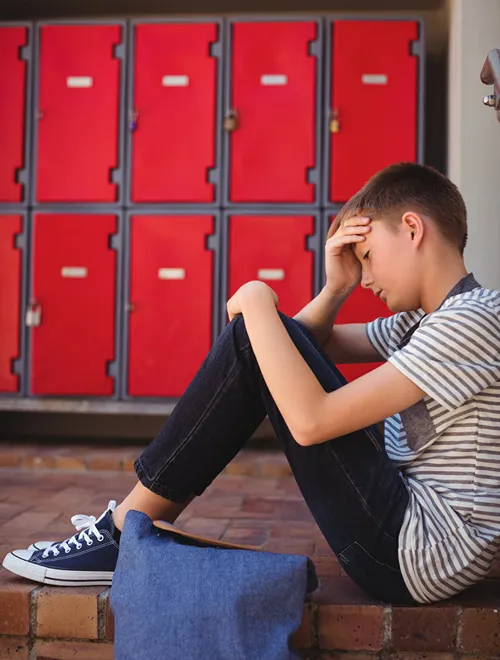 A male teen student sits in front of school locker looking down.