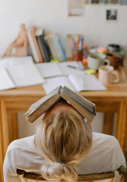 A stress student sitting at a desk looks up places an open book on their face.