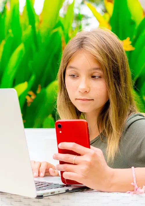 Teen girl typing on her laptop with one hand while holding her smartphone in the other.