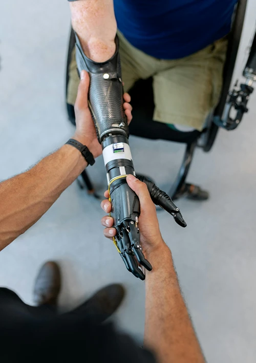 A technician's hands adjusting a carbon-fiber prosthetic arm on a person in a wheelchair, demonstrating a specialized form of assistive technology.