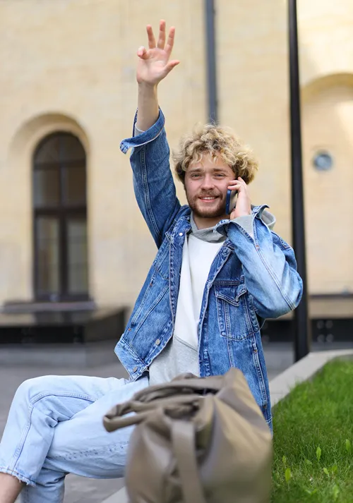 A male student with a travel bag wavs on someone as he talks on his cell phone.