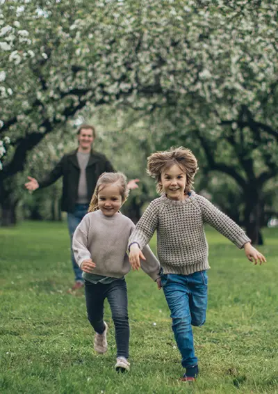 A boy and girl laugh as they run from their dad, who is playfully chasing them in a green meadow with trees.