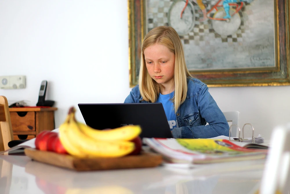 A girl sits at the kitchen table at home doing school work on a laptop.