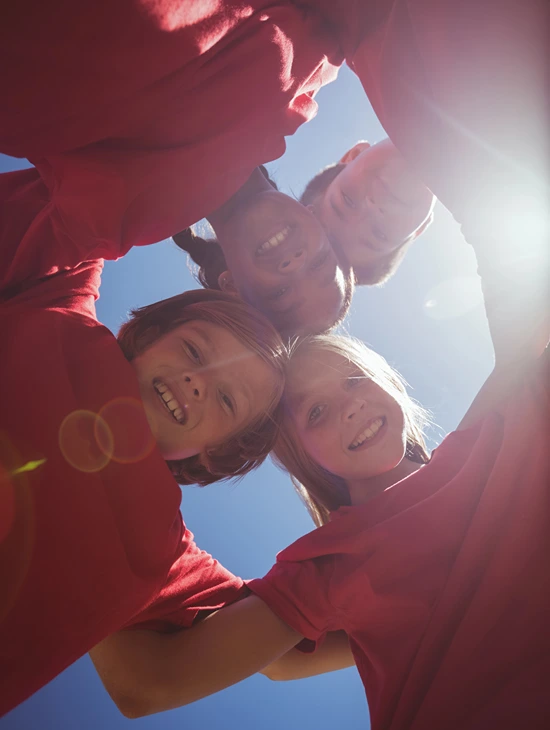 Sun shines down through a group of kids wearing matching t-shirts in a sports team huddle.