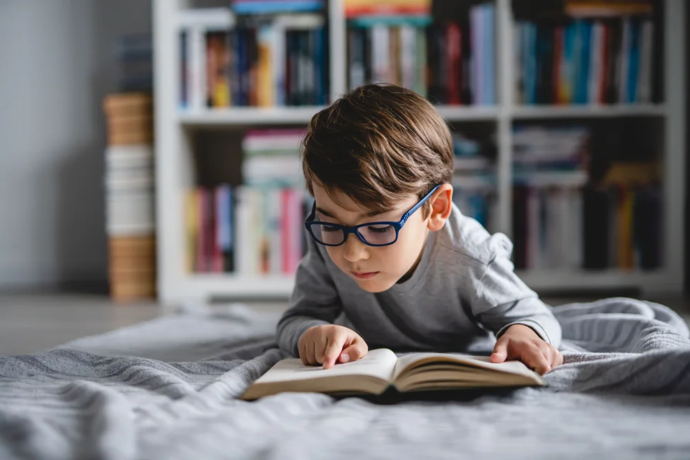 A boy on his bed reading a book with his index finger following each word.