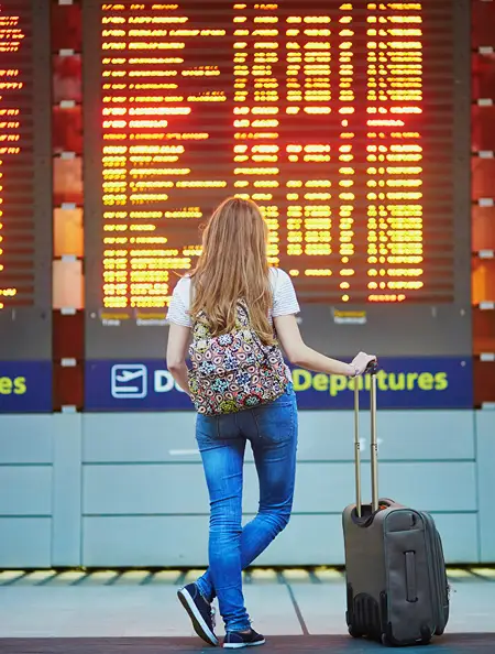 A young woman with long blonde hair, wearing a white striped t-shirt, jeans, and a colorful patterned backpack, stands with her back to the camera holding a rolling suitcase while looking up at a large illuminated flight departures board at an airport.