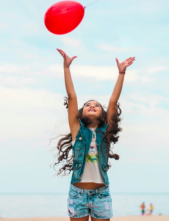 A girl releases a red balloon into the air on a beach.