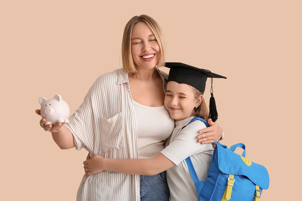 A smiling mother hugs her young daughter, who is wearing a graduation cap and blue backpack, while holding a piggy bank, against a beige background.