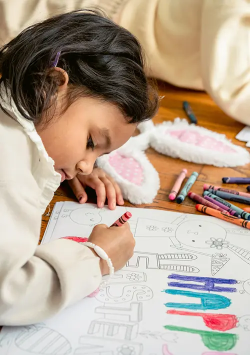 A young girl lying on a wooden floor, coloring in a large coloring book page with a red crayon. Several crayons are scattered nearby, along with a plush bunny toy.
