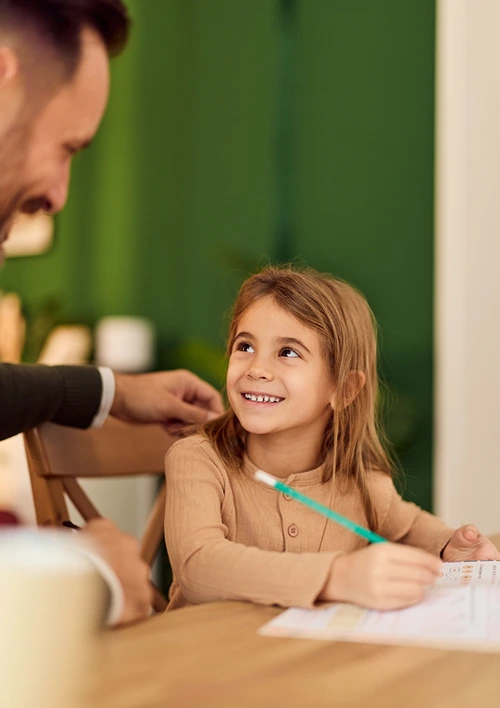A young girl smiles up at her father while doing homework at a wooden table, holding a teal pencil over a worksheet, with a green wall in the background.