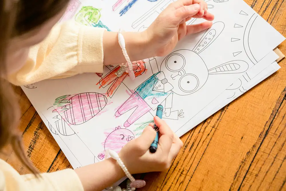 A young girl coloring a large Easter bunny coloring page with crayons on a wooden table