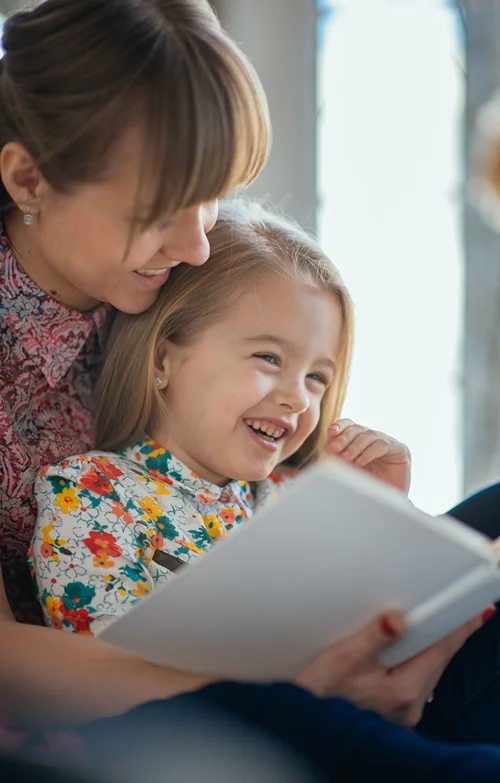 A little girl sits on mom's lap and laughs at an open book.