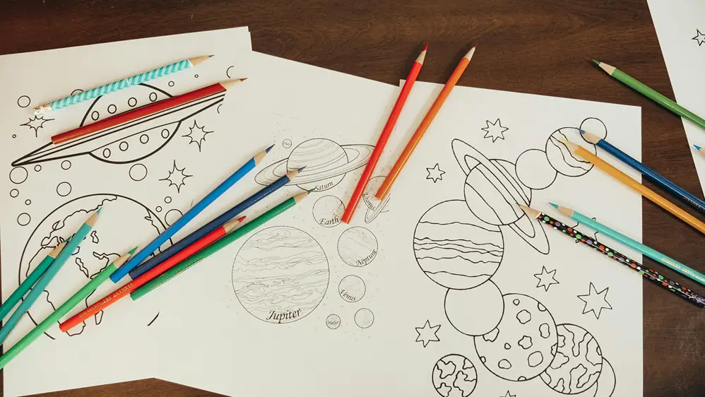 Three uncolored space-themed coloring pages featuring planets, a UFO, and stars, surrounded by colored pencils on a wooden table.