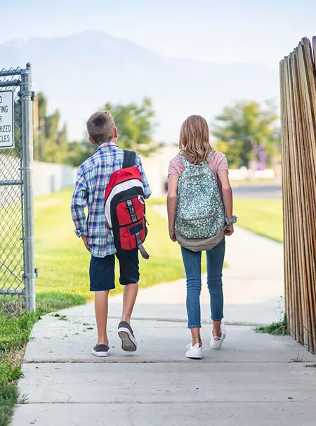 A view from behind of two kids wearing backpacks while walking on a cement pathway.