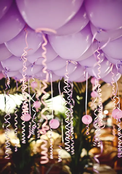Lavender helium balloons with curly pink ribbons and small pink circle weights floating above tropical greenery at an elegant wedding reception decoration.