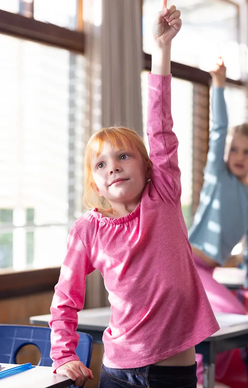 A girl stands at her school desk and stretches to hold up her hand has high as possible.