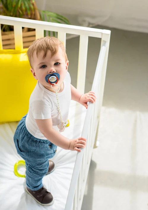 High-angle shot of a toddler with a pacifier in his mouth standing and holding onto the rails.