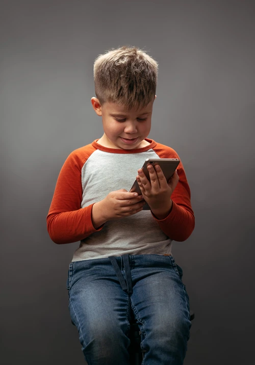 A 12 year old boy looking at a smartphone sits on a stool in a dimly lit room.