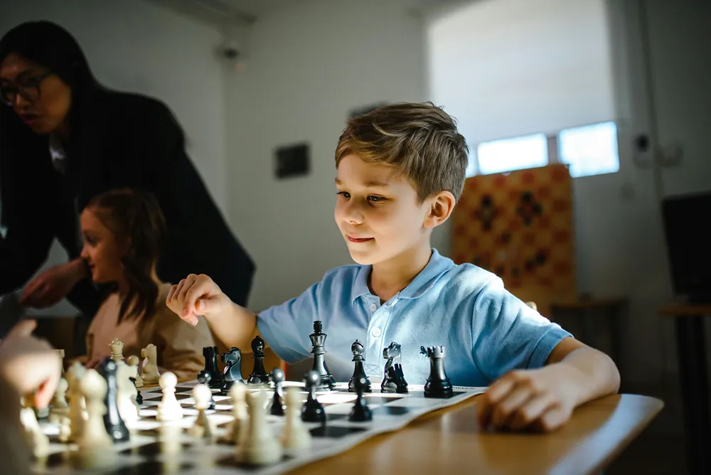 A boy plays chess against an unseen player while a girl in the background play as adult oversees her game.