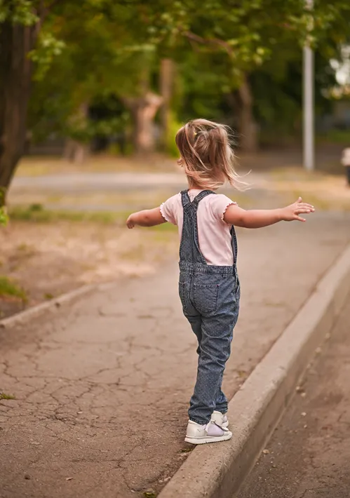 A carefree little girl with her arms outstretched balances and walks on the edge of a sidewalk.