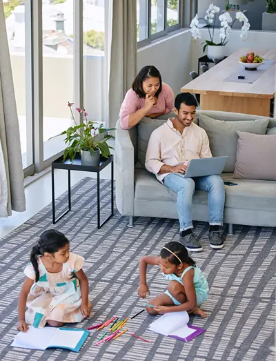 A young family in a bright living room: a man and woman look at a laptop while two young girls draw on a rug.