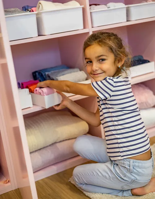 A young girl crouching on the floor, smiling at the camera while organizing folded clothes into white storage bins on a pink shelving unit.