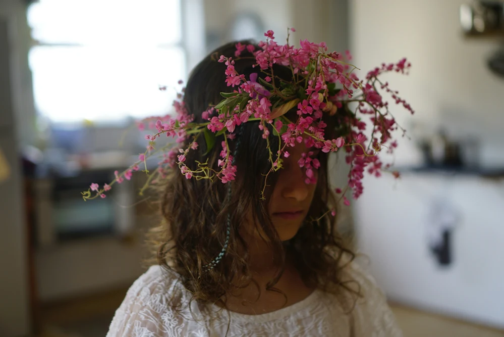 A middle school aged girl with curly brown hair wearing a large, cascading crown of pink wildflowers, head tilted down, dressed in a white lace top with a turquoise necklace, in a softly blurred indoor setting.