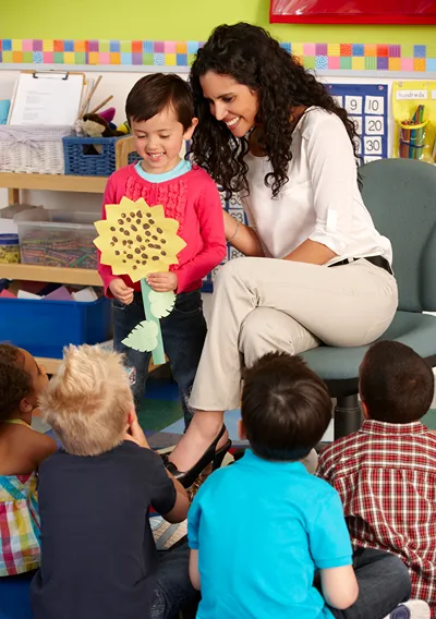 Preschool teacher smiling with a young girl holding a handmade paper sunflower craft in front of her classmates.