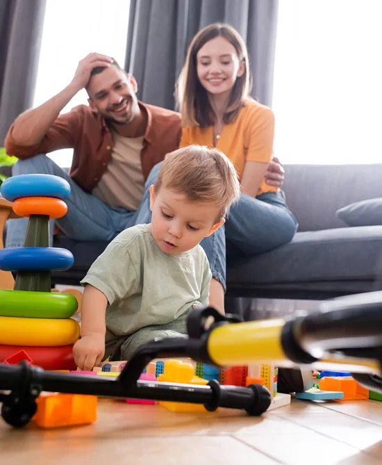 Happy parents sitting on a couch watching their young boy play with toys on a hardwood floor.