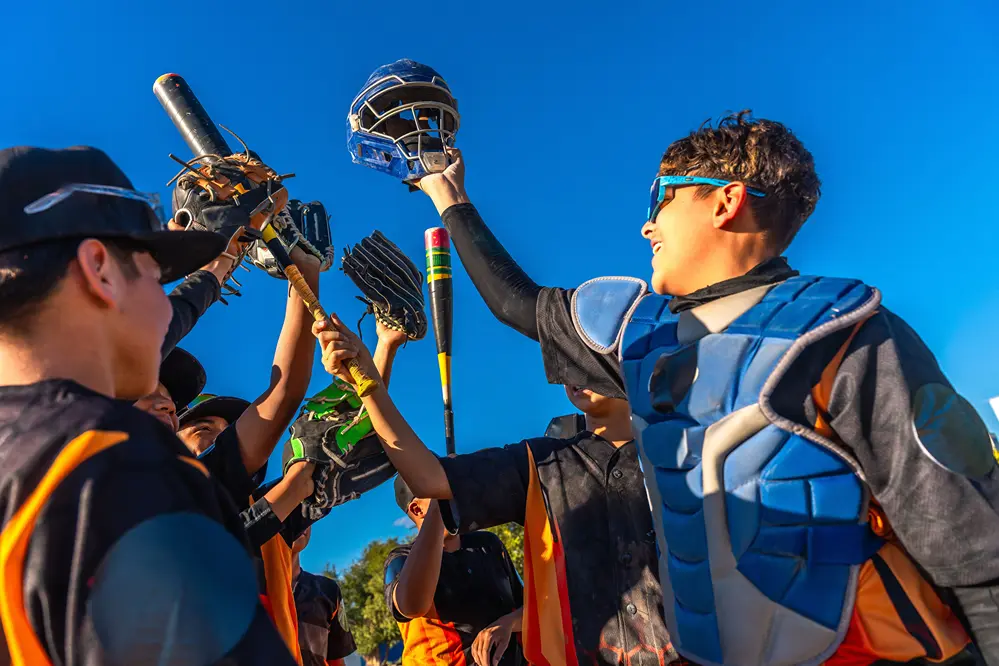 Boys baseball team gather on the field and raise their mitts, bats, and catcher’s facemask together in celebration.
