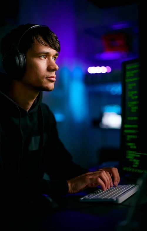 Man in low lit room typing on computer with code on the screen.