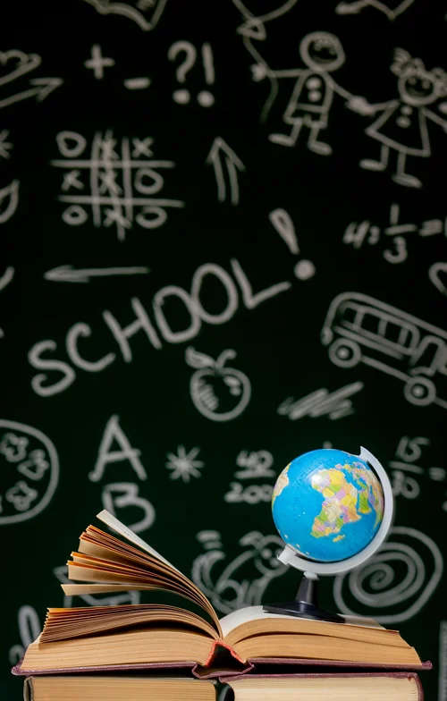 A mini globe sits on an open book on a desk at the front of a classroom. In the background is a blackboard with various symbols and the word SCHOOL written in block letters!