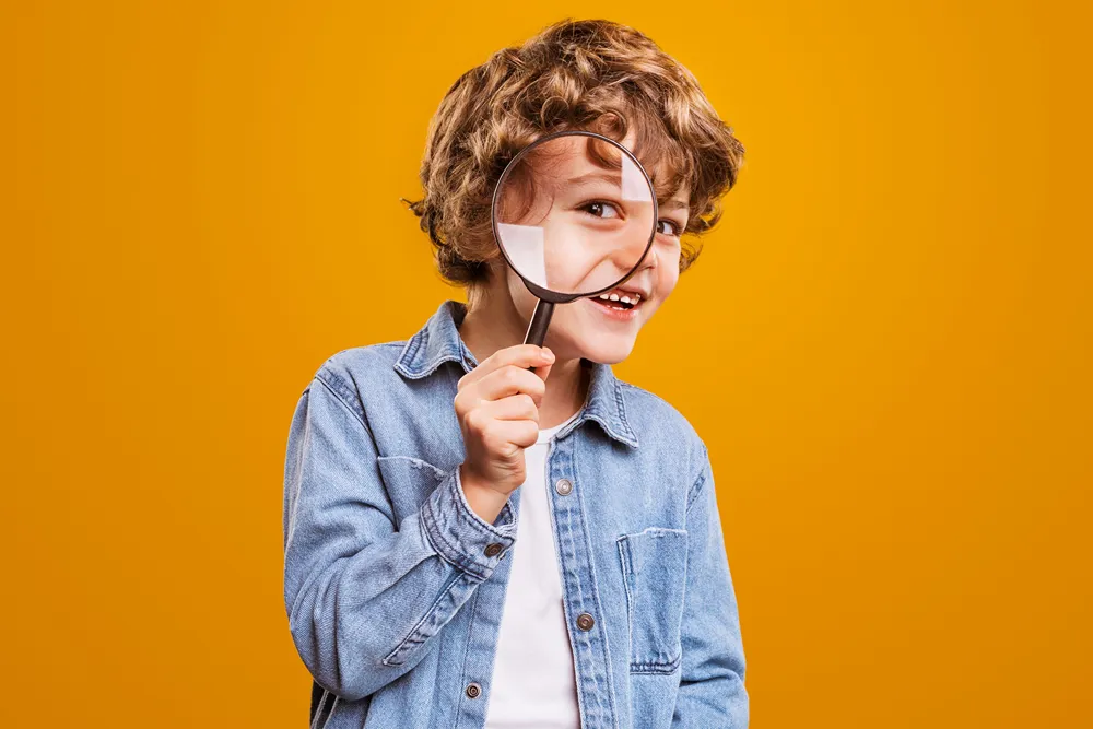 A smiling, curly-haired boy holding a magnifying glass over his eye against a yellow background.
