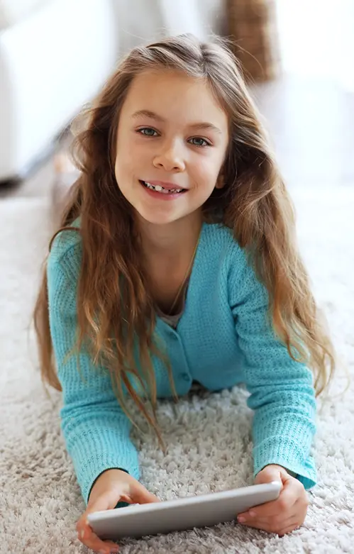 Smiling tween girl on living room floor looks up from playing on her tablet.