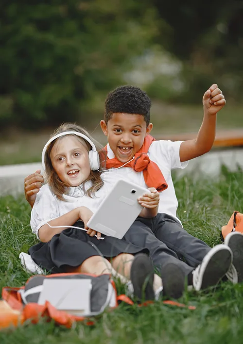 A girl and a boy sitting on the grass in a park with headphones and a tablet.