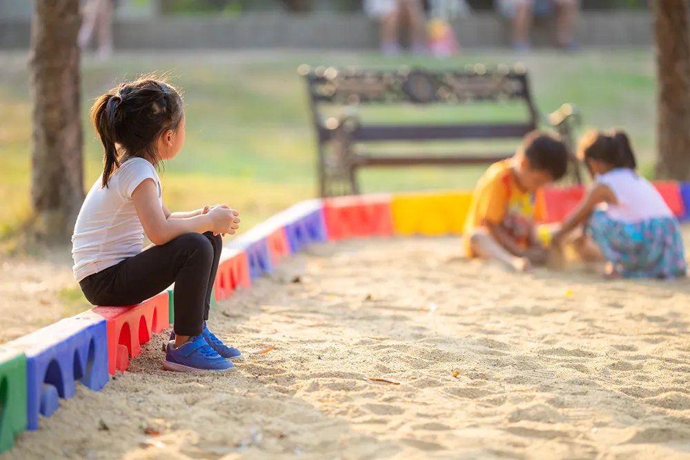 A little girls sits on the edge of a large sand play area watching other kids who play in the distance.
