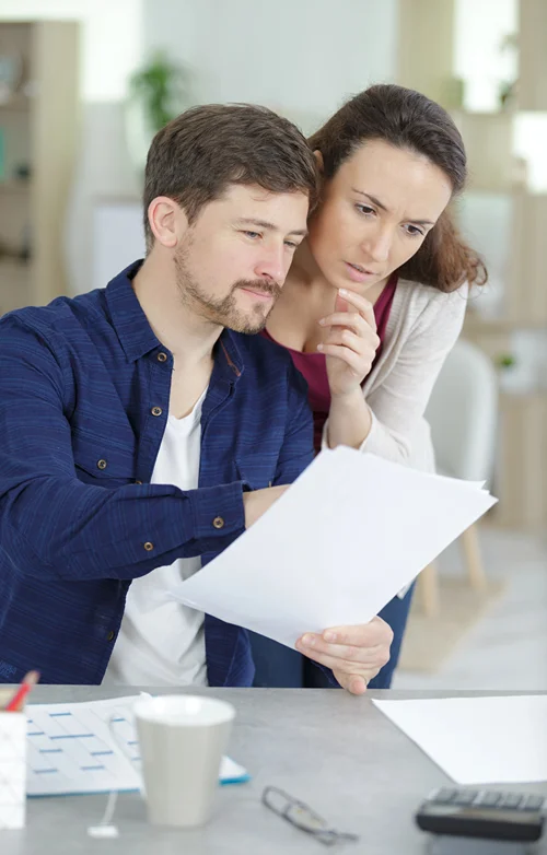 A man sits at a desk with papers and calculator while his wife looks over his shoulder.