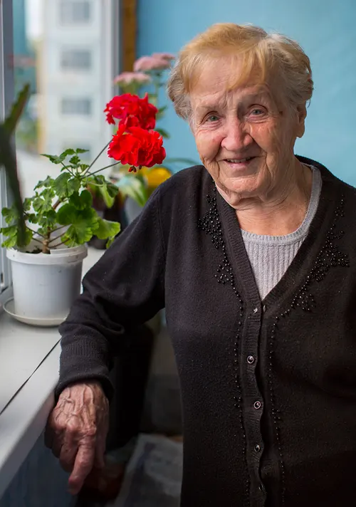 An older woman stands by a flower pot in a window sill.