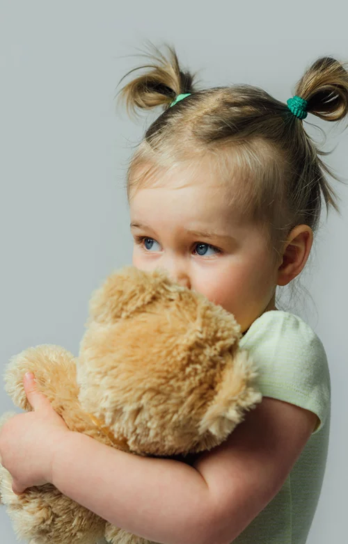 A sheepish girl hides her mouth behind a teddy bear she is holding tightly.
