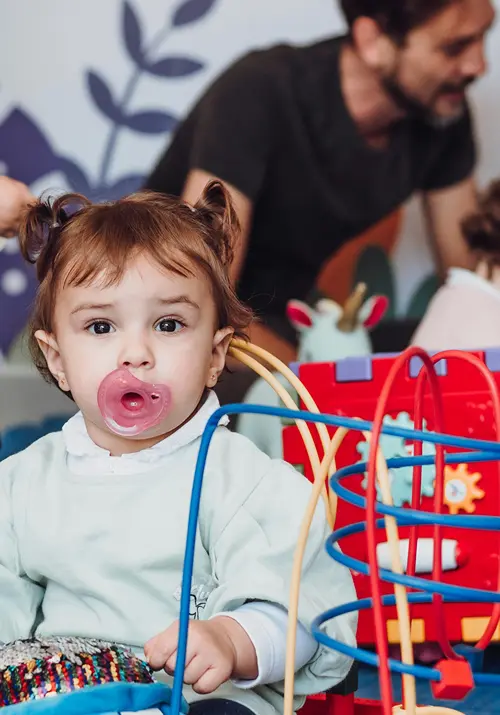 Baby with soother plays with toys in a day care as caregiver plays with other children in the background.