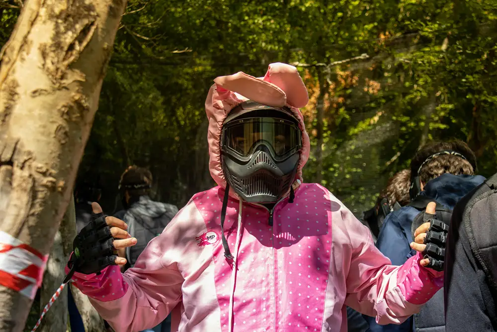 A young person in a pink bunny-eared paintball suit and black mask, standing in a wooded area with other players nearby.