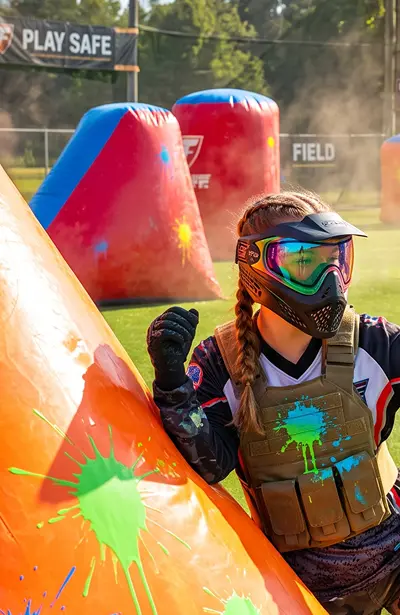 Girl in paintball field leans up against an orange bunker.