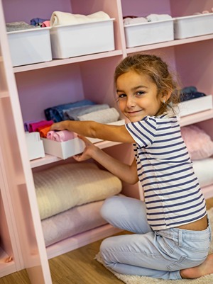 A young girl crouching on the floor, smiling at the camera while organizing folded clothes into white storage bins on a pink shelving unit.