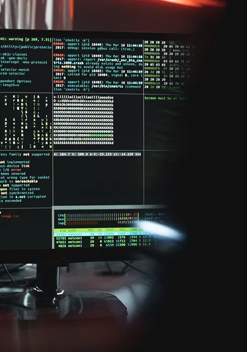 A shadowy figure sits in front of a screen with white and colourful computer code over black.