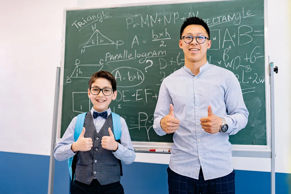 A happy math tutor and elementary student showing success with a thumbs up in front of a chalkboard with math equations.