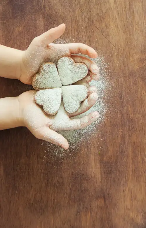 Child's hands holding four heart-shaped cookies dusted with powdered sugar, on a wooden table.